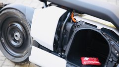 Prtljažnik pod sedežem. Enostaven dostop in uporabna oblika. Za njim se pod belo plastiko skriva elektromotor, baterije so nameščene v dnu skuterja.